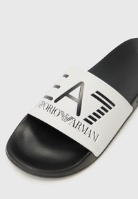 Svart og hvit sklisikker sandal med teksturert såle, med en bred rem prydet med "EA7 Emporio Armani"-logo og horisontale linjer.