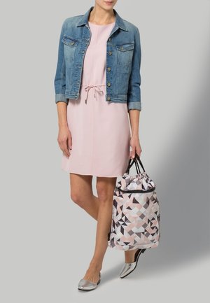 Donna che indossa un vestito rosa e una giacca di jeans blu, porta una borsa con coulisse dal motivo geometrico e indossa scarpe piatte argento.
