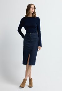 Nainen pukeutuneena tummansiniseen pitkähihaiseen yläosaan, tummansiniseen korkeavyötäröiseen hameeseen, jossa on edessä halkio, ja vaaleanruskeisiin nilkkureihin, seisomassa valkoista taustaa vasten.