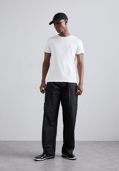 T-shirt blanc à manches courtes avec un petit logo, associé à un pantalon cargo noir avec des poches latérales et des chaussures noires à la finition brillante.
