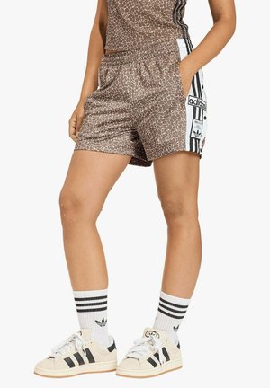 Persona che indossa pantaloncini sportivi marroni con motivo a righe e pannelli laterali bianchi con loghi Adidas, abbinati a calzini bianchi Adidas e scarpe da ginnastica beige.