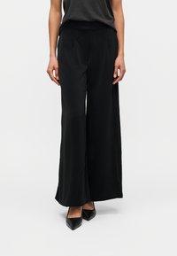 Γυναίκα που φοράει μαύρο παντελόνι με φαρδύ μπατζάκι, μαύρες μυτερές γόβες και μαύρο γκρι αμάνικο τοπ που είναι μαζεμένο μέσα στο παντελόνι, στέκεται μπροστά σε ομοιόμορφο ανοιχτό φόντο.