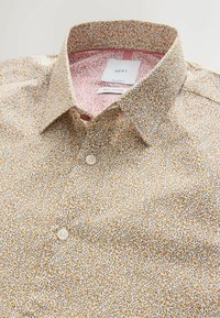 Chemise en coton avec un fond crème et un motif floral jaune et marron. Elle présente un col classique et des boutons blancs.