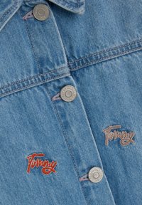 Tela de mezclilla azul con botones plateados etiquetados "Tommy Hilfiger NYC" y dos logotipos bordados de "Tommy" en hilo rojo y beige.
