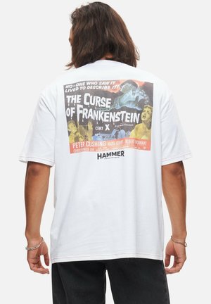 T-shirt en coton blanc avec un imprimé d'affiche de film vintage au dos, avec des graphiques colorés et du texte, à manches courtes et à coupe décontractée.