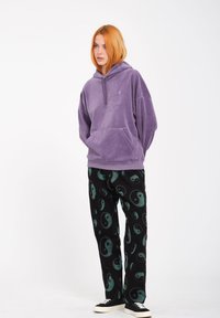Felpa viola in velluto con tasca, abbinata a pantaloni neri con motivi yin-yang verdi e scarpe nere.