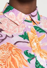 Chemise à manches courtes à boutons avec un imprimé floral, fond lavande, grandes fleurs oranges et roses, et feuilles vertes, dotée d’un col classique.