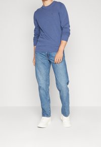 Homme portant un pull en tricot à manches longues bleu, un jean bleu clair et des baskets blanches, debout devant un fond clair uni.
