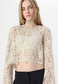 Top court en dentelle crème avec un motif floral, manches longues, encolure ronde et ourlet brut. Associé à un pantalon sombre et ajusté.