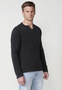 Juodas ilgomis rankovėmis marškinėliai su henley apykakle, tekstūruota medžiaga ir subtiliu logotipo detaliu krūtinėje, derinami su šviesiai mėlynomis džinsais.