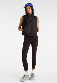 Svart vadderad väst med hög krage, dragkedjestängning, bärs över åtsittande svarta leggings. Blå keps med logotyp, grå sportskor och vita strumpor.