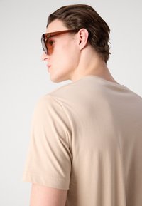 Beige bomulds t-shirt med korte ærmer, vist fra ryggen, med en glat tekstur og en rund halsudskæring. Mørke solbriller fremhæver looket.