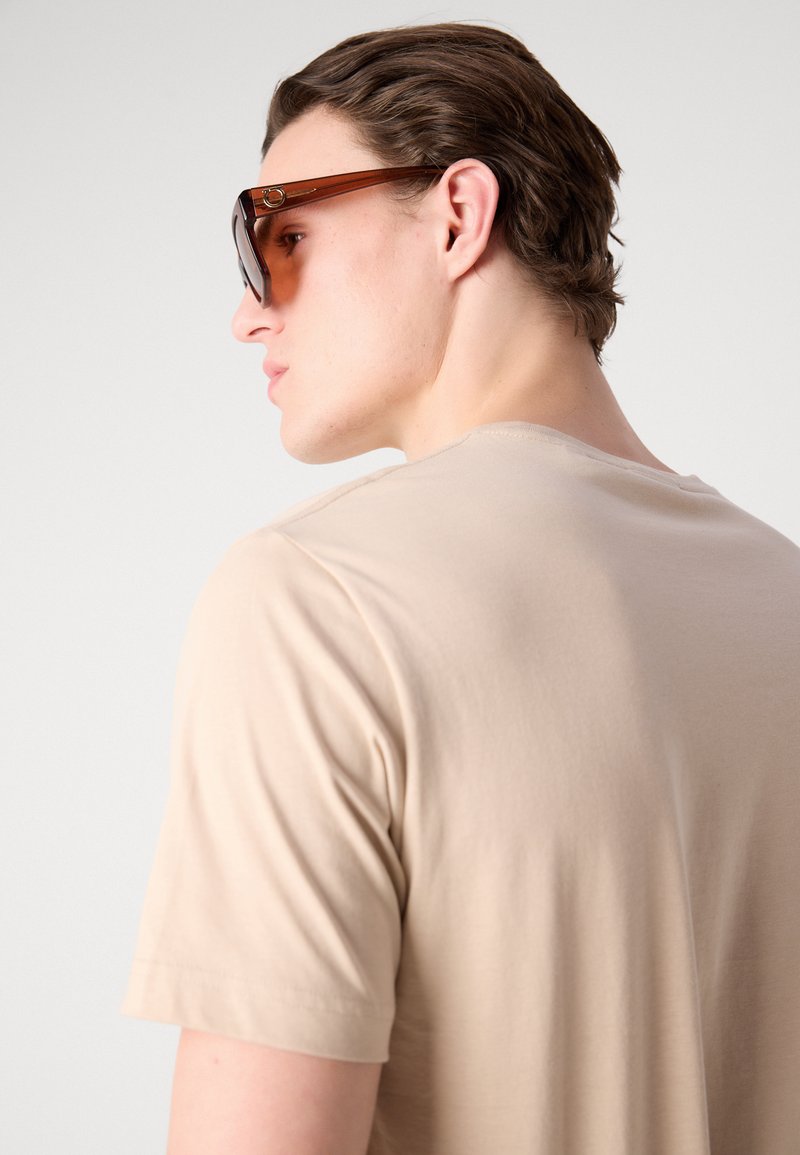 Beige Baumwoll-T-Shirt mit kurzen Ärmeln, von hinten gezeigt, mit einer glatten Textur und einem runden Ausschnitt. Dunkle Sonnenbrillen vervollständigen den Look.