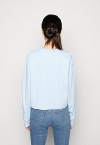 Donna con capelli scuri raccolti in un chignon, vestita con una felpa azzurro chiaro e jeans blu, in piedi di fronte a un muro leggero e uniforme.