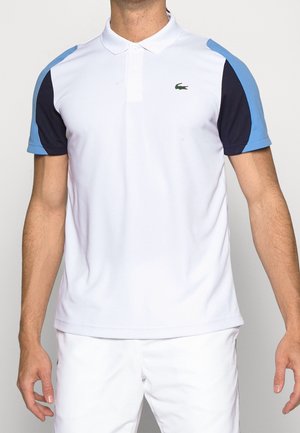 Uomo che indossa una polo bianca con maniche a blocchi di colore azzurro chiaro e blu navy e piccolo logo Lacoste verde sul petto.