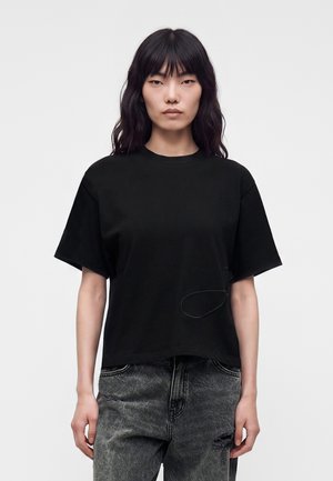 Femme aux longs cheveux noirs portant un t-shirt noir uni à manches courtes et un jean noir délavé, debout devant un fond uni.