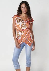 Blusa blanca con estampado floral y acentos en naranja y amarillo, con mangas cortas con volantes y un diseño cruzado. Combinada con unos vaqueros recortados en azul claro con dobladillos decorados.