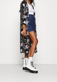 Lang sort blomster-kimono med pink og hvide blomster, båret over en hvid tanktop og blå denimshorts. Hvide snørebåndsstøvler med sorte såler.
