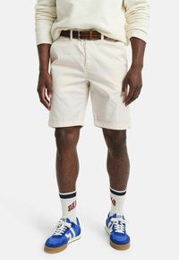 Lichtbeige shorts met opgerolde zomen, gedragen met een bruin gevlochten riem. Gecombineerd met blauwe sneakers en crèmekleurige sokken met marineblauwe en rode accenten.