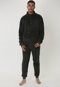 Ung man som står barfota, iförd en mörkgrön fluffig fleece-tröja och matchande jogger-byxor mot en enkel bakgrund.