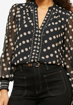 Zwarte doorschijnende blouse met lange mouwen en beige polkadots, knoopsluiting aan de voorkant, een getand randje, gecombineerd met zwarte hoge taille broek en riem.