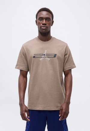 Άνδρας που κοιτάζει εμπρός, φοράει καφέ μπλουζάκι με μαύρο και λευκό λογότυπο μπάσκετ και μπλε σορτς, σε απλό λευκό φόντο.