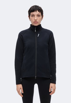 Personne aux cheveux courts et foncés portant une veste noire zippée et un pantalon noir, debout devant un fond clair et uni.