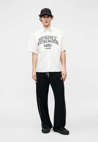 Junger Mann trägt ein weißes "SPRINT SUMMER 2026 MSGM"-Shirt, schwarze weite Hosen, schwarze Mütze und schwarze Sneakers, steht vor einem einfarbigen Hintergrund.