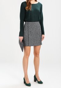 Groene geribbelde sweater met lange mouwen, zwart-wit patroon rok, groene suède hoge hakken, met een grijze clutches.