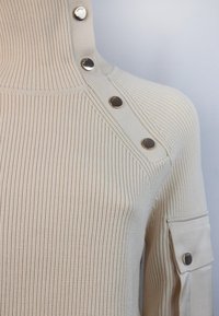 Pull en maille côtelée crème avec col montant. Présente des boutons-pression métalliques sur l'épaule et une poche avec détail bouton sur la manche.