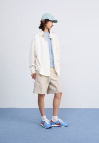 Camicia oversized crema chiaro sopra una maglietta azzurro chiaro, pantaloni corti beige e sneakers blu con accenti arancioni; sfondo azzurro chiaro.