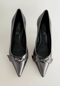 Fekete lakkbőr magassarkúk hegyes orral és csomózott masni díszítéssel elöl, sima felülettel és párnázott talppal.