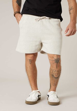 Homme portant un short blanc à cordon, un t-shirt noir, des baskets blanches, avec des tatouages sur les deux jambes et des bracelets au poignet gauche.