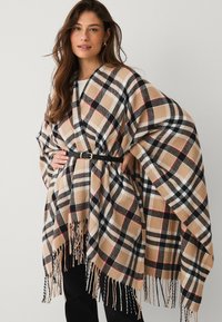 Plaid poncho in beige, black, and red tones. Features fringed edges and a black belt at the waist for a structured look. Soft texture.