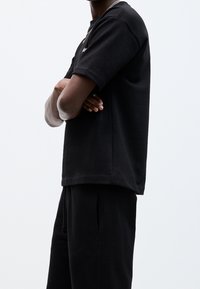 Personne portant une chemise noire à manches courtes et un pantalon noir, bras croisés, visible des épaules à la taille sur un fond uni.