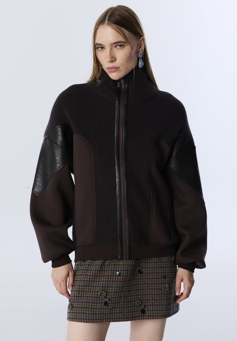 Vrouw in een donkerbruine hoodie met rits, voorzien van leren panelen en een hoge kraag, gecombineerd met een geruite minirok versierd met kleine kraaltjes.