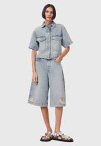 Giacca di jeans azzurro chiaro con maniche corte e orlo borchiato, abbinata a pantaloni corti azzurro chiaro dal fit morbido e sandali metallici. Presenti dettagli stropicciati.