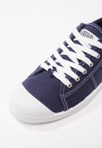 Scarpa in tela blu navy con punta in gomma bianca, lacci bianchi e un piccolo dettaglio decorativo vicino ai lacci. Design classico di sneaker basse.