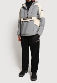 Grå och gräddfärgad puffjacka med halvdragkedja, logodetalj och justerbar huva, kombinerad med svarta fleecebyxor och mörka kängor.