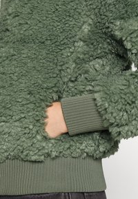Grön, texturerad fleecejacka med en mjuk yta, ribbade manschetter och fåll, med en sidoficka integrerad i tygets design.