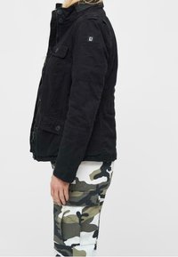 Giubbotto di jeans nero con zip frontale, bottoni e tasche a patta. Indossato con pantaloni cargo camouflage bianchi e verdi.