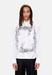 Άτομο με μακριά dreadlocks που φοράει λευκό πουκάμισο με κουμπιά, διακοσμημένο μπροστά με κυκλικό μαύρο αφηρημένο σχέδιο που μοιάζει με αγκάθια.