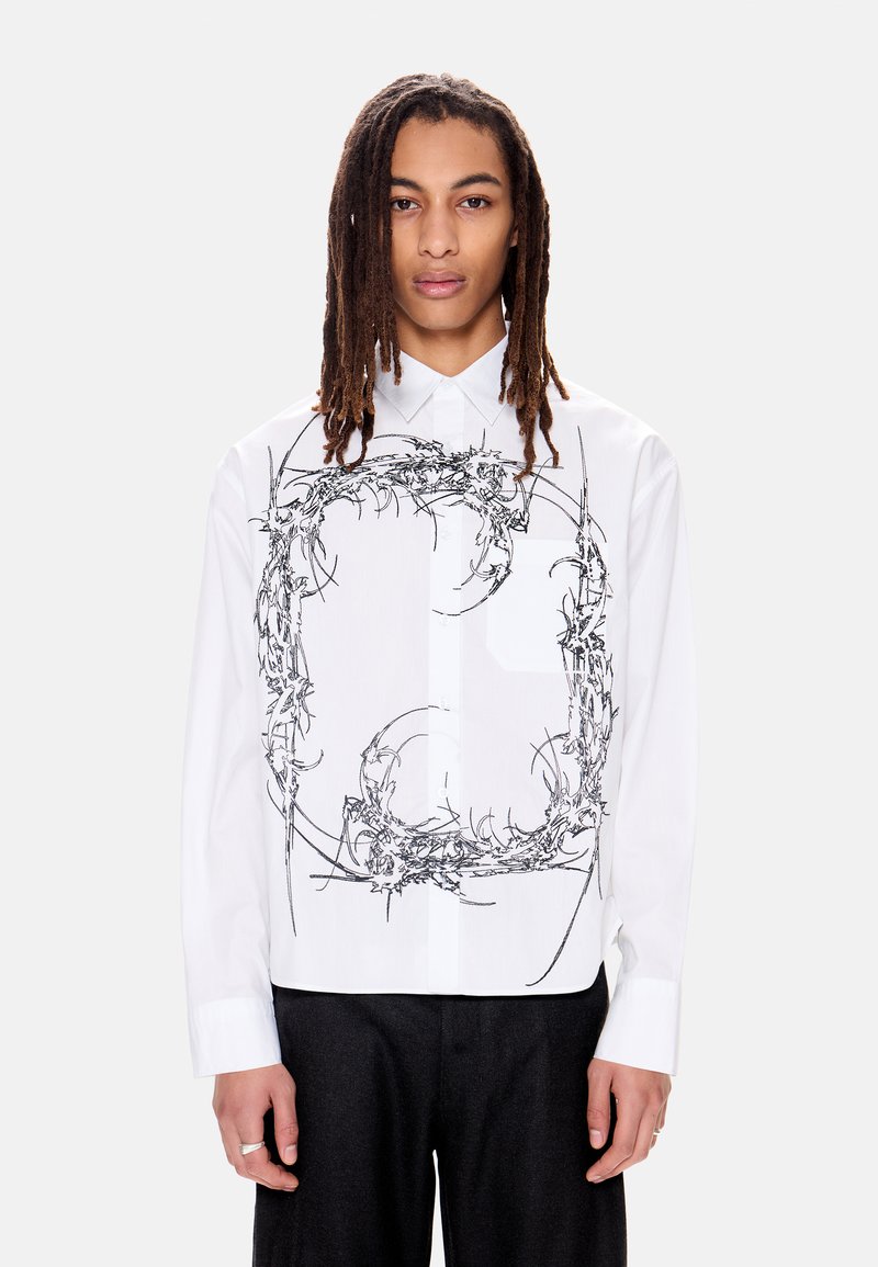 Άτομο με μακριά dreadlocks που φοράει λευκό πουκάμισο με κουμπιά, διακοσμημένο μπροστά με κυκλικό μαύρο αφηρημένο σχέδιο που μοιάζει με αγκάθια.