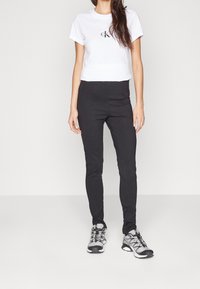 Weißes Baumwoll-T-Shirt mit kleinem schwarzem Logo, kombiniert mit schmalen schwarzen Hosen und grauen Sportschuhen mit schwarzen Akzenten und Schnürsenkeln.