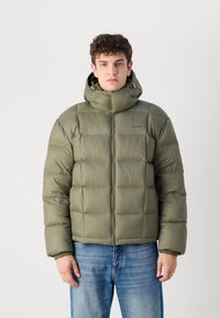Polstret jakke i olivengrønn, med horisontal quiltet tekstur, høy krage og frontglidelås. Brukt med lyseblå jeans.