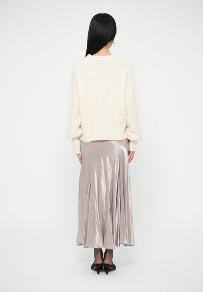 Světlý krémový svetřík s kabelovým vzorem v kombinaci s dlouhou, lesklou svetle šedou saténovou maxi sukní. Oblečení má dlouhé rukávy a volný střih.