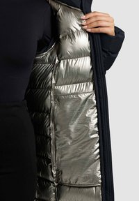 Mujer sosteniendo un abrigo negro abierto con un forro interior acolchado metálico plateado brillante y un bolsillo interior.
