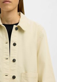 Jasnobeżowa kurtka wykonana z bawełny, z kołnierzem, czarnymi guzikami i kieszenią na piersi. Tkanina ma subtelną fakturę i czysty design.