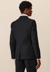 Giacca nera da uomo vista da dietro, con spacco singolo, taglio sartoriale, polsini con bottoni e colletto di camicia bianca visibile sotto.