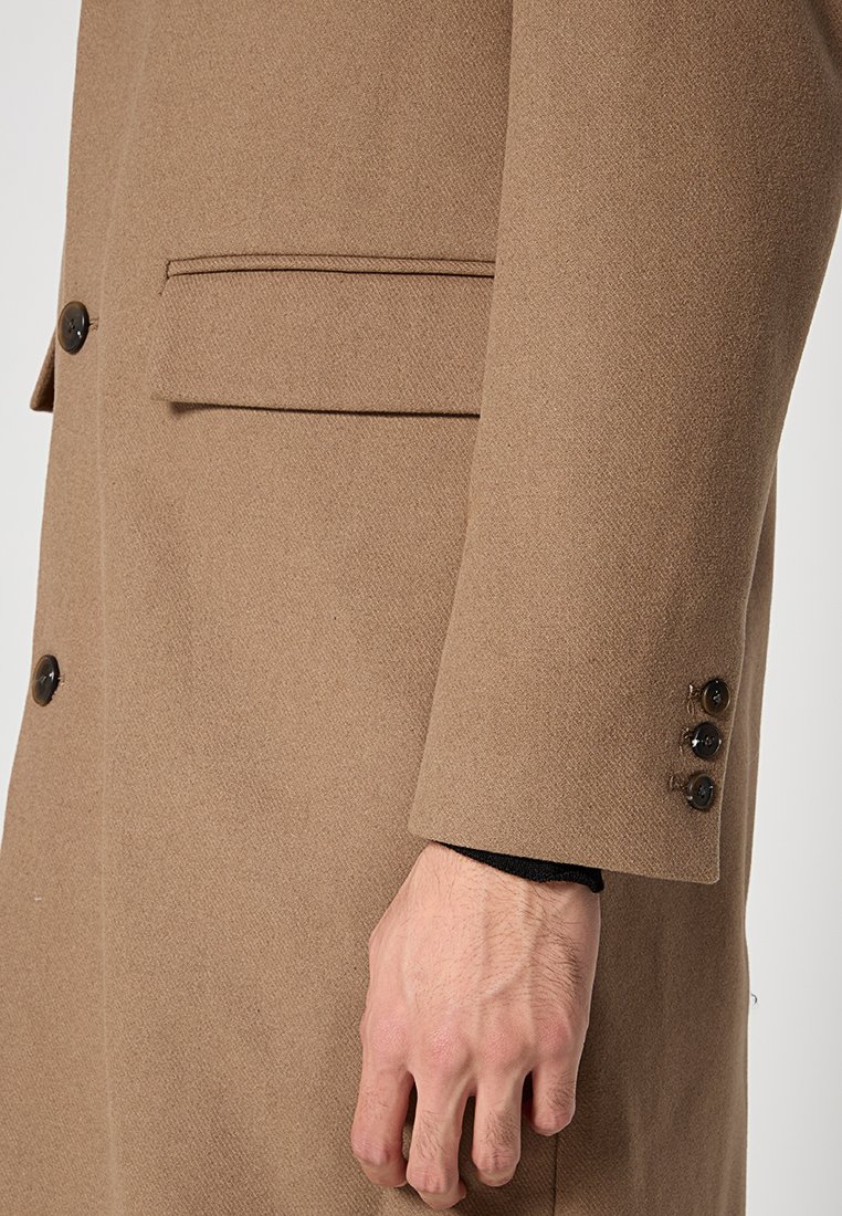 Cappotto in lana marrone con una tasca sul petto, dettagli con tre bottoni ai polsini e una vestibilità sartoriale. La texture appare morbida e calda.